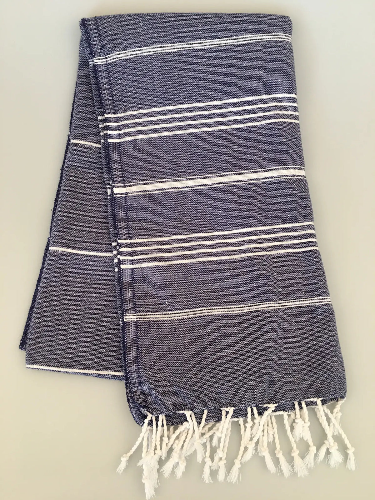 "Dark blue peshtemal towel with white stripes and fringe edges, neatly folded on a light background."