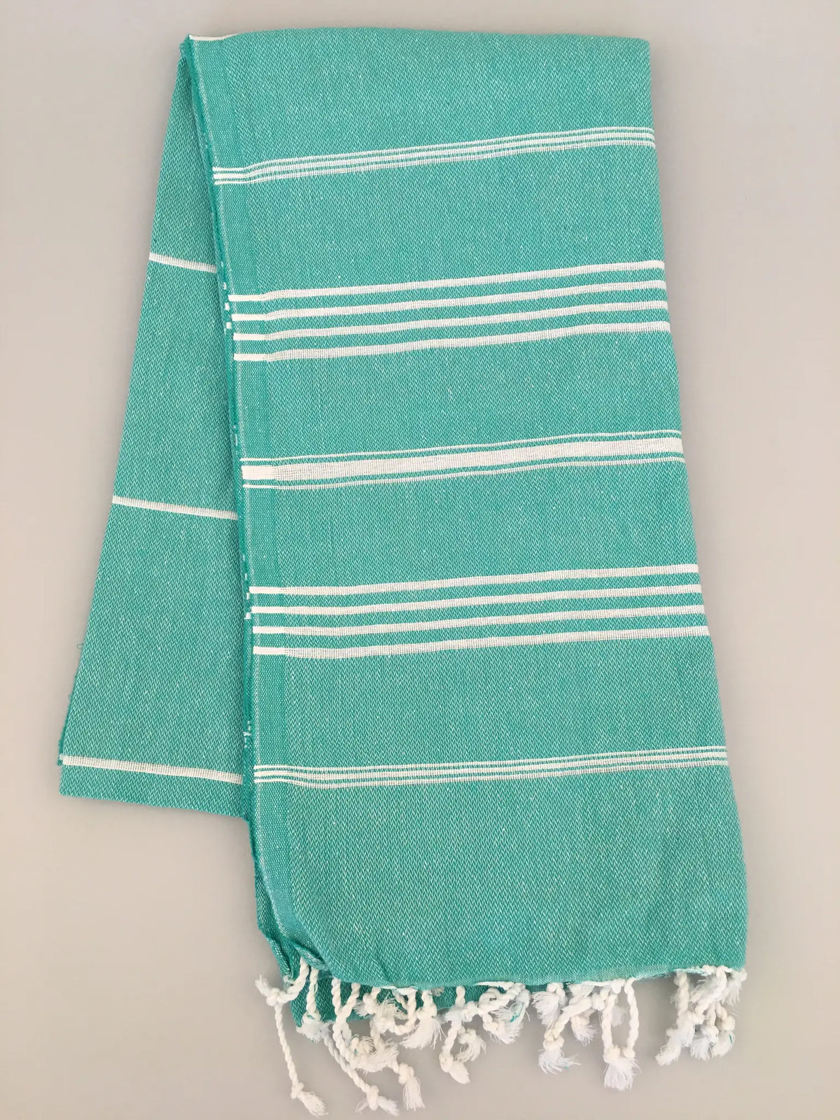 Turquoise Turkish towel with white stripes and fringed ends, laid flat on a light gray background.