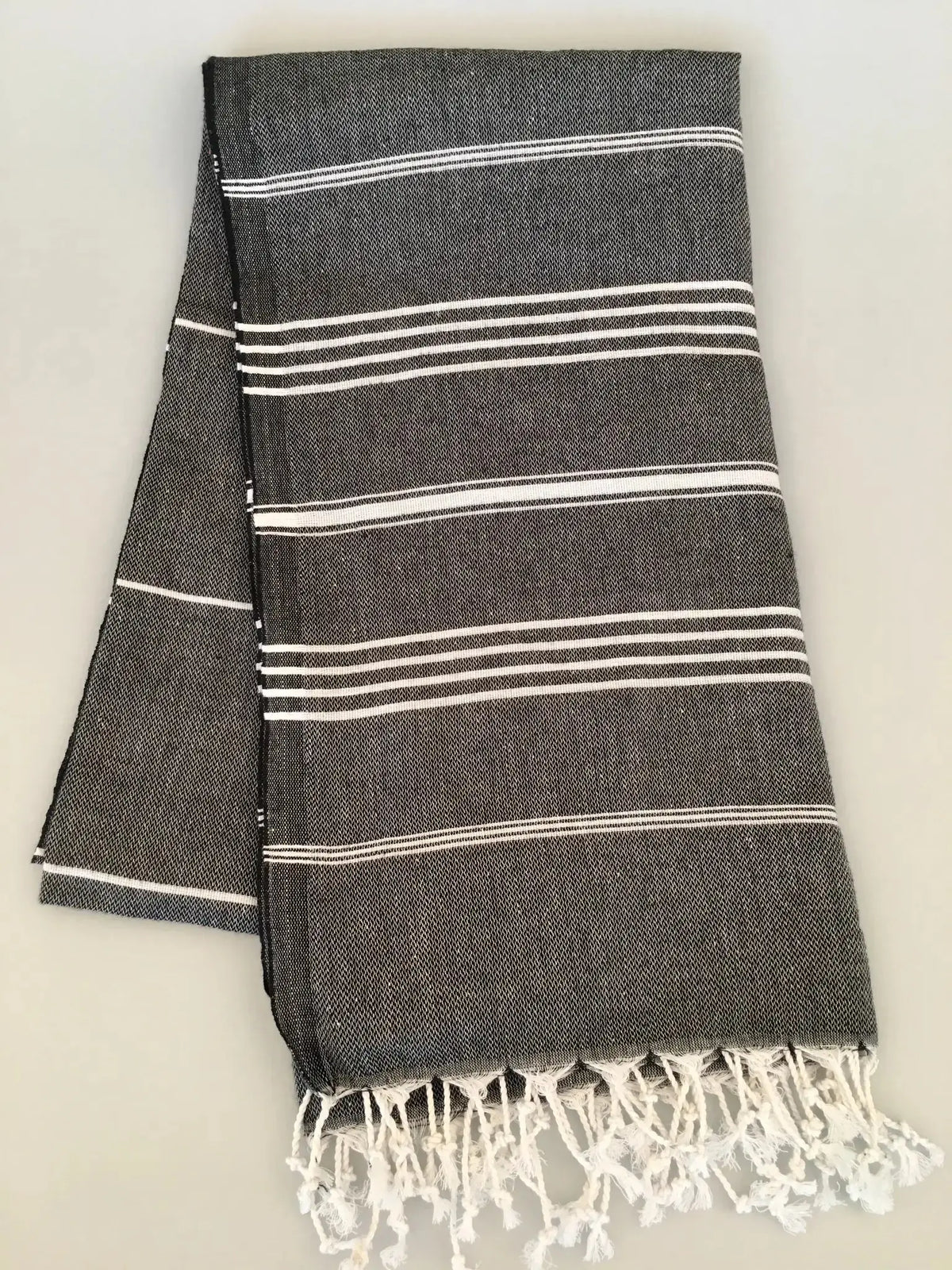 "Dark gray Turkish towel with white stripes and fringed edges, folded flat on a light background."